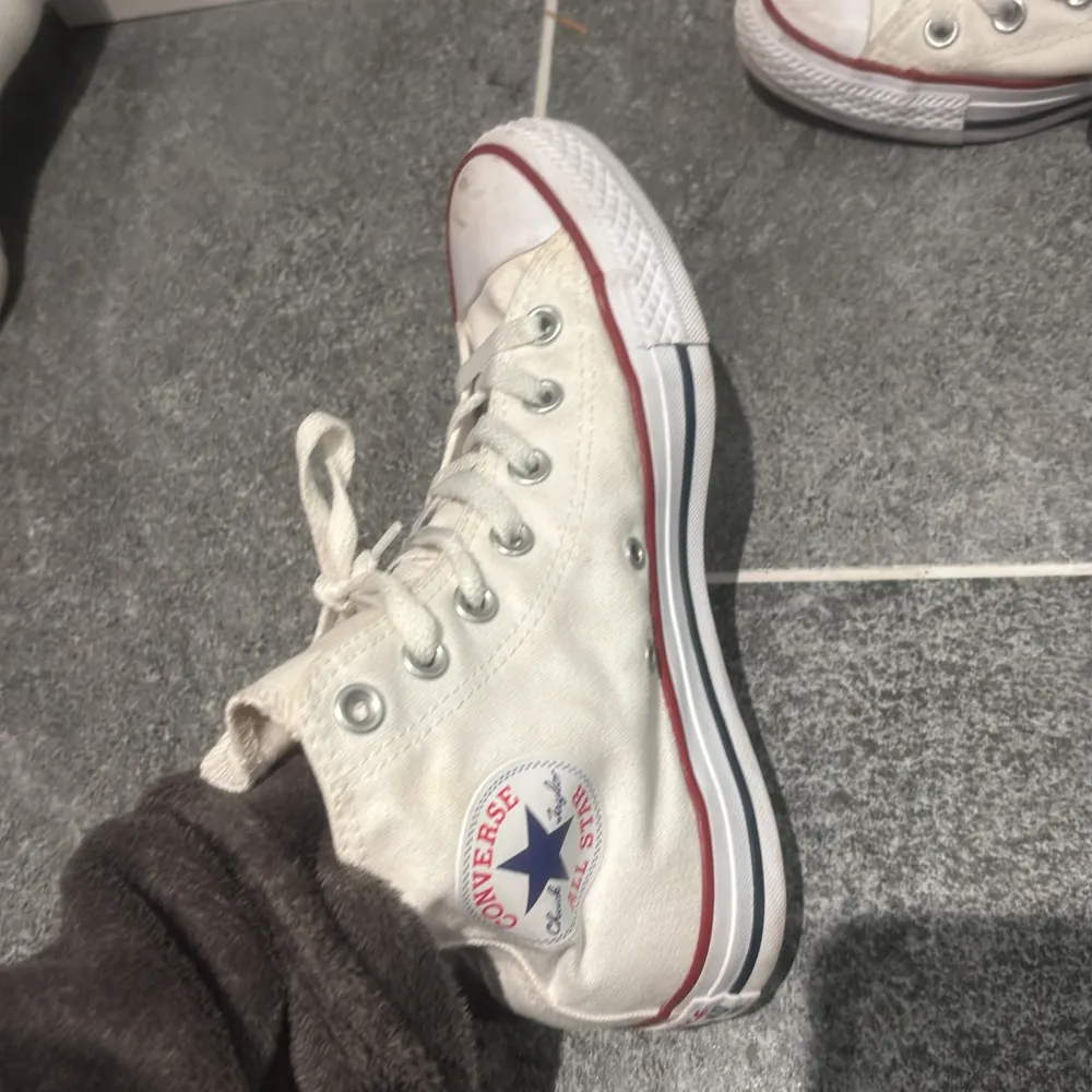 Säljer ett par klassiska vita Converse All Star i bra skick. De har den ikoniska höga modellen med snörning och röd/blå rand runt sulan. Perfekta för både vardag och fest! Lite slitna men fortfarande snygga och bekväma.. Skor.