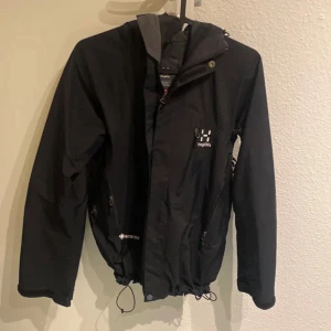 Svart vindjacka från Haglöfs - Säljer en svart vindjacka från Haglöfs i bra skick. Jackan har dragkedja och knappar framtill, samt justerbar huva. Perfekt för höst och vår med sitt slitstarka material och stilrena design. Passar både till vardags och utomhusaktiviteter. Pris kan diskuteras 