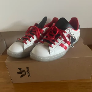Suuupersnygga sneakers!! - Säljer ett par snygga Adidas sneakers i vitt och rött. De är adidas campus x Howlin rays. Jättebra skick endast använda fåtal gånger. Vita skosnören och den svarta hängande detaljen medföljer. Storlek 40! Skriv om ni har frågor💕