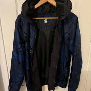 Blå camo vindjacka från G-Star RAW - Säljer en snygg blå camo vindjacka från G-Star RAW. Jackan har en dragkedja framtill och en praktisk huva. Perfekt för höst och vår, med ett coolt mönster som sticker ut. Passar bra till både vardag och lättare utomhusaktiviteter.