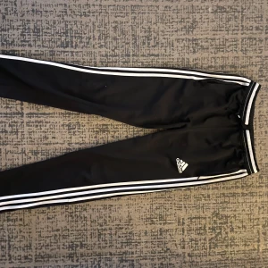 Svarta träningsbyxor från Adidas - Säljer ett par svarta träningsbyxor från Adidas. Byxorna har de klassiska tre vita ränderna längs sidorna och en elastisk midja med snörning. Perfekta för träning eller chill. Materialet är lätt och andas bra, vilket gör dem sköna att ha på sig under längre perioder.