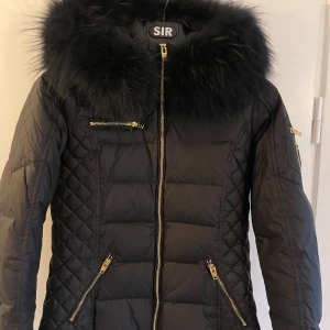 Svart quiltad jacka med pälsdetalj från Rock and Blue - Snygg svart quiltad jacka från SIR med fluffig pälsdetalj på huvan. Jackan har dragkedjor i guld och flera praktiska fickor. Perfekt för kyliga dagar och ger en lyxig känsla. Passar både till vardags och fest!