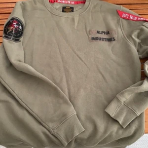 Olivgrön sweatshirt från Alpha Industries - Säljer en olivgrön sweatshirt från Alpha Industries i mycket bra skick. Den har långa ärmar och coola detaljer som en patch på ärmen och en röd etikett. Perfekt för höst och vår!