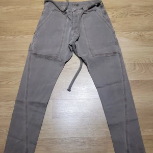 Grå byxor från SixtyDays - Säljer ett par grå byxor från SixtyDays i storlek XS. De har en avslappnad stil med knytband i midjan och stora fickor med knappar. Perfekta för en casual look och tillverkade i Portugal. Passar bra till både vardag och avslappnade tillfällen.