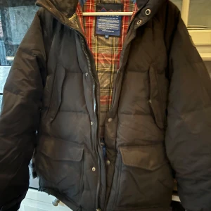 Svart dunjacka från Penfield - Säljer en svart dunjacka från Penfield i storlek M. Jackan har en klassisk rutig insida i rött och grått, och är perfekt för kalla dagar. Den har en dragkedja och knappar framtill för extra skydd mot kylan. Perfekt för vintern! 🖤