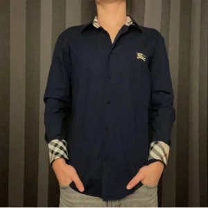 Mörkblå skjorta från Burberry - Säljer en snygg mörkblå Burberry-skjorta i slim fit. Den har långa ärmar och klassiska Burberry-rutiga detaljer på insidan av kragen och ärmsluten. Passar M då den är slimfit! Perfekt för både vardag och fest! 🕺