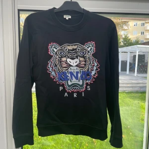 Svart sweatshirt från Kenzo - Säljer en svart sweatshirt från Kenzo med ett coolt tigertryck i flera färger på framsidan. Tröjan har långa ärmar och en normal passform. Perfekt för höst och vår när du vill ha något stilrent men ändå något i ögonfallande. 