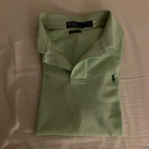 Grön pikétröja från Ralph Lauren - Säljer en snygg grön pikétröja från Ralph Lauren. Den har en klassisk krage och korta ärmar, perfekt för både vardag och lite finare tillfällen. Tröjan är i bra skick och har den ikoniska loggan broderad på bröstet. Passar perfekt till jeans eller chinos för en stilren look.