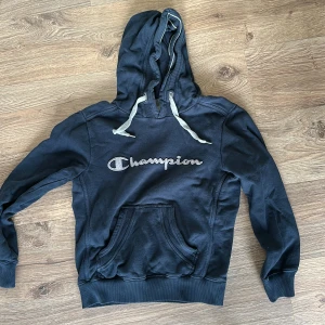 Svart hoodie från Champion - Säljer en mörkblå hoodie från Champion i bra skick. Den har en klassisk känguruficka och justerbar huva med vita snören. Perfekt för en avslappnad stil eller träning. Passar både höst och vår.