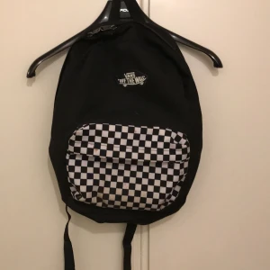 Svart och vit ryggsäck från Vans - Säljer en cool svart ryggsäck från Vans med klassiskt schackrutigt mönster på fickan. Perfekt för skolan eller vardagsbruk. Ryggsäcken har justerbara remmar och en praktisk dragkedja. Den är i bra skick och redo för nya äventyr!