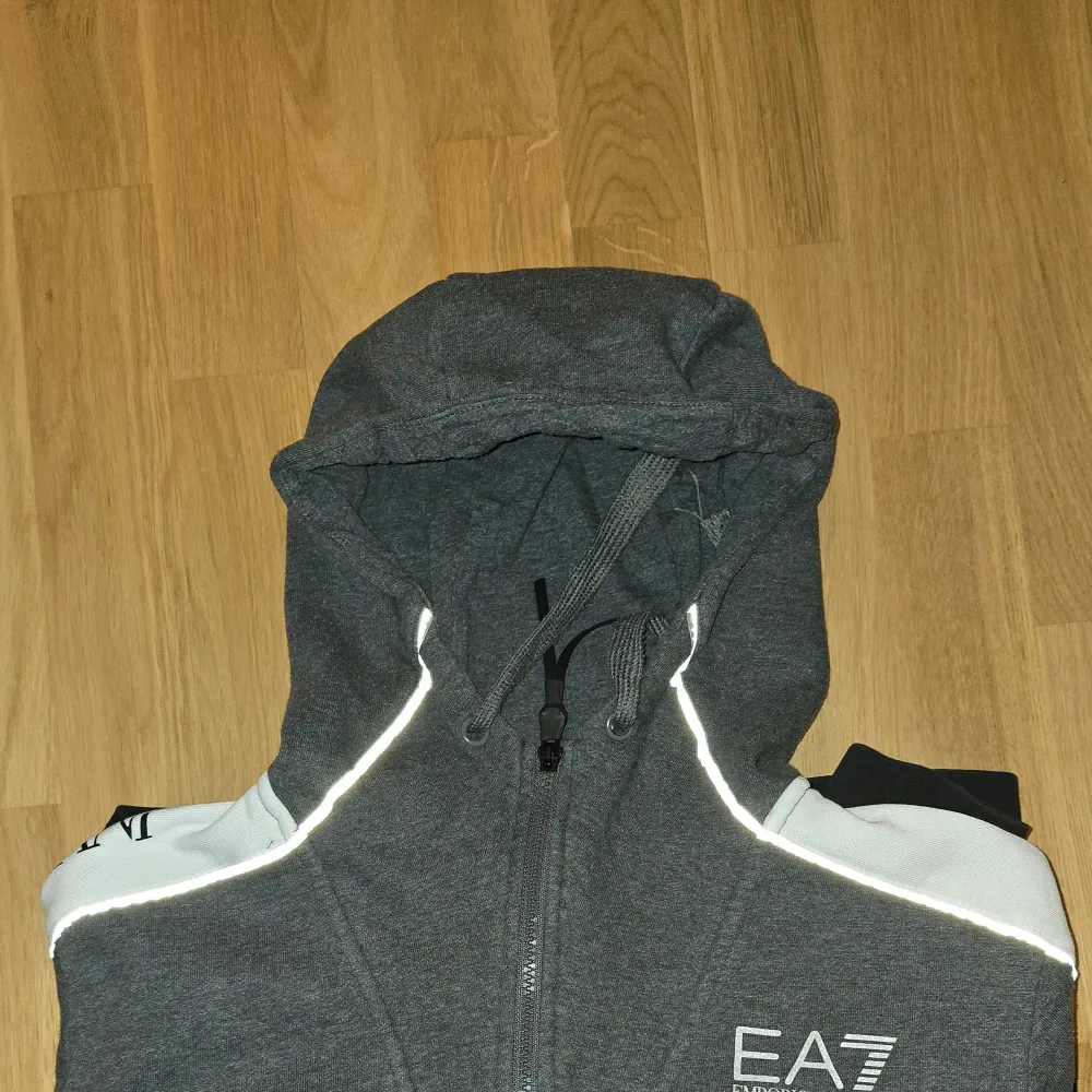 Säljer en snygg grå och svart hoodie från Emporio Armani. Den har en dragkedja framtill och en stor luva. Ärmarna är dekorerade med vita detaljer och loggan EA7 tryckt på bröstet och ärmen. Perfekt för en casual look eller träning. Hör av dig vid intresse! . Hoodies.