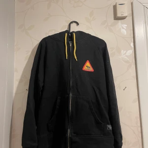 Svart hoodie med Norland-tryck - Säljer en svart hoodie med dragkedja och gult snöre i luvan. Den har ett coolt Norland-tryck på ryggen och en liten logga fram. Perfekt för höst och vinter, och den är i bra skick. Stor ficka fram för att hålla händerna varma. Passar både till vardags och chillkvällar. Ena fickan har gått upp lite grann se bild 3 men går att fixa om man kan sy övrigt bra skick!