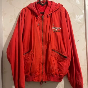 Röd vintage jacka med tryck - Säljer en cool röd vintage jacka med tryck på både fram- och baksidan. Jackan har en huva som är avtagbar och dragkedja framtill, samt praktiska fickor med dragkedjor. Perfekt för en avslappnad stil och att sticka ut i mängden!