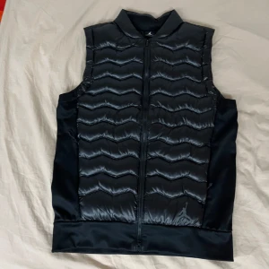 Svart dunväst från Jordan - Säljer en snygg svart dunväst från Jordan i storlek M. Västen har en quiltad design med dragkedja framtill och det ikoniska Jordan-logotypen på bröstet. Perfekt för höst och vår när du vill hålla dig varm men ändå se stilren ut. Dessutom så är den aldrig använd med lapp kvar på.