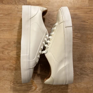Vita sneakers från Zara - Säljer ett par stilrena vita sneakers från Zara i nytt skick. De har en klassisk design med snörning och är perfekta för både vardag och fest. Skorna är gjorda i syntetmaterial och har en bekväm passform. Perfekta för vår och sommar! 