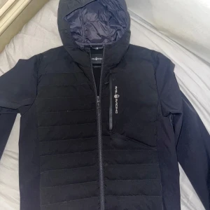 Svart hybrid jacka från Sail Racing  - Säljer en svart jacka från Sail Racing i storlek L. Den har en stilren design med dragkedja och huva. Perfekt för höst och vår, med quiltade detaljer som ger extra värme. Jackan är i mycket bra skick och passar både till vardags och lite mer sportiga aktiviteter.