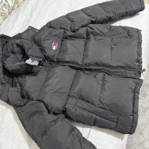 Svart dunjacka från Tommy Hilfiger - Säljer en snygg svart dunjacka från Tommy Hilfiger. Jackan har en dragkedja framtill och en praktisk huva. Perfekt för kalla vinterdagar och håller dig varm och stilren. Den är i mycket bra skick och redo för nya äventyr!