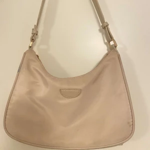 Beige axelväska med dragkedja - Säljer en stilren beige axelväska i syntetmaterial. Väskan har en praktisk dragkedja upptill och en justerbar axelrem. Perfekt för vardagsbruk med en neutral färg som passar till allt. Några små fläckar finns på sidan, men annars i bra skick.