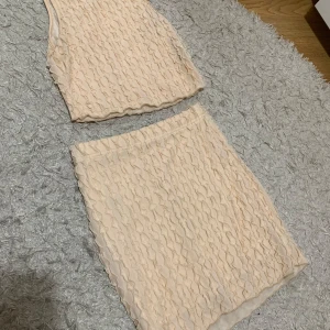 Beige ärmlös topp och kjol - Säljer ett set med en beige ärmlös topp och matchande kjol. Båda delarna har ett unikt volangmönster som ger en elegant touch. Perfekt för sommarens fester eller en dag på stan. Toppen är i ett mjukt och bekvämt material.