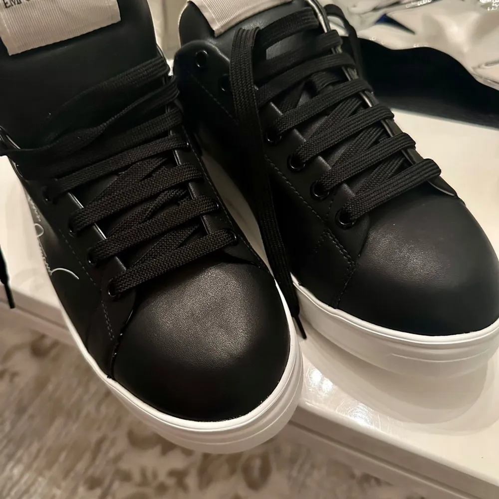 Snygga och äkta svarta sneakers från Emporio Armani med vit sula och vit signaturdetalj på sidan. Ny pris 3000 kr. Skorna har en stilren design med svart snörning och en bekväm passform. Perfekta för både vardag och festliga tillfällen! Storlek 43. Köpta från zalando. Priset kan diskuteras. Kvitto finns. Säljer eftersom dom passade inte.. Skor.