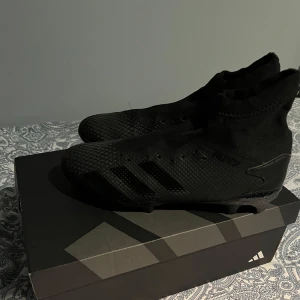 Svarta fotbollsskor från Adidas - Säljer ett par svarta fotbollsskor från Adidas i mycket bra skick. Skorna har en strumpliknande passform och är perfekta för spel på gräs. De har en snygg, helsvart design med diskreta ränder på sidan. Perfekta för dig som vill dominera på planen med stil! Storlek 41
