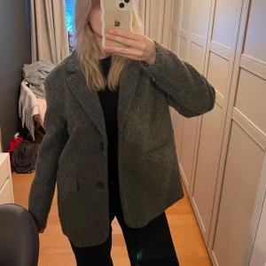 Grå ullkavaj - Säljer en snygg grå kavaj i ullmaterial. Den har en klassisk design med knappar framtill och långa ärmar. Perfekt för höst och vinter, och funkar både till vardags och fest. Storleken är normal och den är i mycket bra skick. Använd 1 gång 