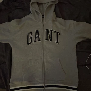 Grå hoodie från GANT - Säljer en snygg grå hoodie från GANT i bra skick. Den har en dragkedja framtill och stora bokstäver på bröstet. Perfekt för höst och vår, med ribbade muddar och en skön huva. Passar både till vardags och chillkvällar. 🧥