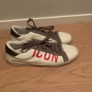 Vita sneakers från Dsquared2 - Säljer ett par vita sneakers från Dsquared2 med texten 'ICON' i rött på sidan. Skorna har bruna/gråa snören och detaljer. Perfekta för en casual look och passar till både jeans och shorts. Använda men i bra skick. Har tyvärr inte kvar boxen. Skick 8/10.