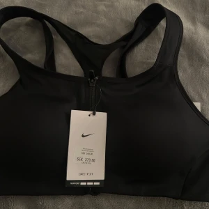 Svart sport-bh från Nike - Säljer en ny svart sport-bh från Nike med Dri-FIT-teknologi. Den har en stilren design med brottarrygg och ett elastiskt band under bysten för extra stöd. Perfekt för träning och andra aktiviteter. Prislappen sitter kvar.