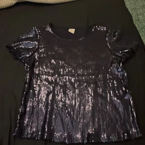 Svart glittrig topp från Lindex - Säljer en svart glittrig topp från Lindex i storlek 134/140 men passar även för 12-14 åringar. Perfekt för festliga tillfällen med sitt glittrande paljettmaterial. Toppen har korta ärmar och en bekväm passform. Perfekt för att lysa upp vilken outfit som helst!