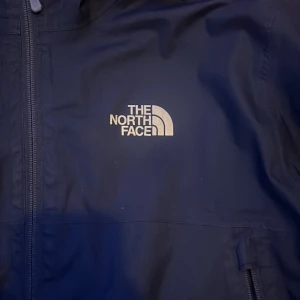 Svart vindjacka från The North Face - Säljer en svart vindjacka från The North Face i bra skick. Jackan har en huva och dragkedja framtill, perfekt för blåsiga dagar. Den är stilren och funktionell med två praktiska fickor med dragkedja. Passar både till vardags och utomhusaktiviteter.