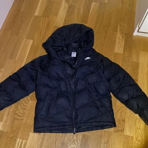 Barn storlek svart pufferjacka från Nike - Säljer en svart pufferjacka från Nike i mycket bra skick. Jackan har en dragkedja och en huva, perfekt för kalla dagar. Den är tillverkad i syntetmaterial och passar bra för både höst och vinter. Superbekväm och stilren design! Skick 9/10 finns ingen syn av andvändning