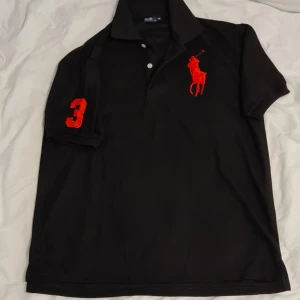 Svart pikétröja från Ralph Lauren - Säljer en snygg svart pikétröja från Ralph Lauren med röd broderad logga på bröstet och siffran 3 på ärmen. Perfekt för en stilren och sportig look. Tröjan har en klassisk krage och knappar framtill. Passar både till vardags och fest!