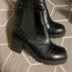 Svarta boots med krokodilmönster - Snygga svarta boots i skinn med krokodilmönster och klack. De har en dragkedja på sidan för enkel på- och avtagning. Perfekta för höst och vinter, dessa boots ger en cool touch till vilken outfit som helst.