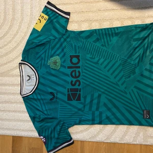 Newcastle borta tröja. Storlek M.  - Säljer en grön fotbollströja med ett coolt geometriskt mönster. Tröjan har korta ärmar och en rund halsringning med svart och vit kant. Märket Sela syns tydligt på framsidan. Perfekt för fotbollsfans som vill sticka ut på planen!