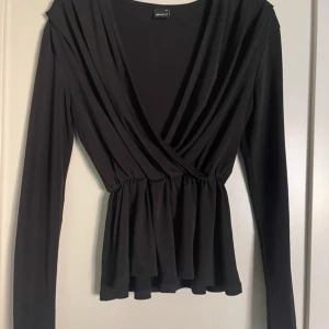 Svart blus från Gina - Snygg svart omlottopp från Gina i mjukt material. Den har långa ärmar och en smickrande v-ringning. Perfekt för både vardag och fest! 🖤