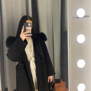Svart kappa med pälsdetaljer - Säljer en elegant svart kappa med fluffig pälsluva (äkta päls). Perfekt för att hålla sig varm och stilren under kyliga dagar. Den har en klassisk design med långa ärmar och en bekväm passform. Passar perfekt för både vardag och festliga tillfällen. Köpt för ca 2700kr på femenina säljes för 2000kr kan skickas omgående och väldigt bra skick!