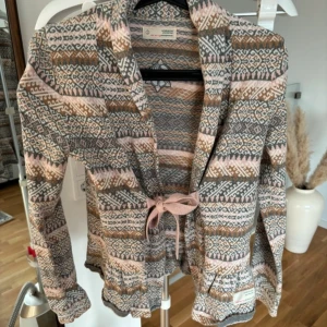 Mönstrad kofta från Odd Molly strl 1 - Superfin mönstrad kofta från Odd Molly i mjuka toner av rosa, brunt och grått. Den har långa ärmar och knytband framtill för en söt look. Perfekt för höst och vår när du vill ha något mysigt och stiligt. Passar både till vardags och lite finare tillfällen.