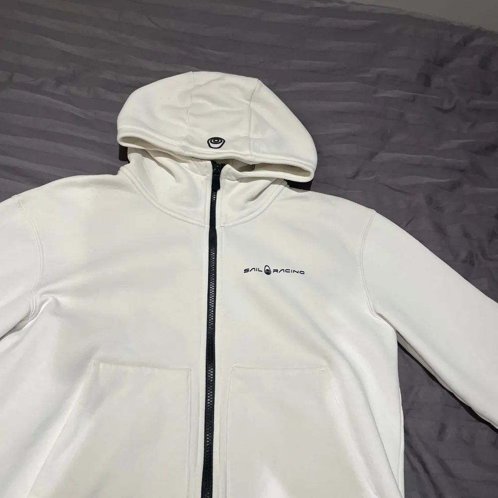 Säljer en snygg vit hoodie från Sail Racing i mycket bra skick. Den har en dragkedja framtill och en praktisk huva. Perfekt för både vår och höst. Materialet känns mjukt och skönt, och den har en stilren design med logotypen på bröstet.. Tröjor & Koftor.