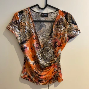 Vintage topp - Snygg och färgglad omlottopp från L'Onkel i ett härligt mönster med orange, svart och beige toner. Toppen har en v-ringad design och korta ärmar, perfekt för en somrig look. Materialet känns mjukt och följsamt, vilket gör den bekväm att bära hela dagen. Str m