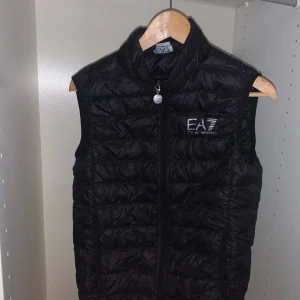 Svart  EA7 - Snygg svart dunväst från EA7 i mycket bra skick. Perfekt för höst och vår, med dragkedja framtill och quiltad design. Västen har en stilren look och är idealisk för både vardag och fest. Passar perfekt över en hoodie eller tröja för extra värme.