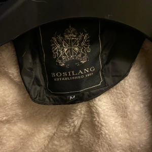 Svart jacka från Bosilang - Säljer en svart jacka från Bosilang i storlek M. Jackan har en quiltad design med en mysig, ljus insida som ser ut att vara i fleece. Perfekt för kyliga dagar med sin långa ärmar och dragkedja framtill. Den har också en snygg krage med knappar.