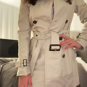 Beige trenchcoat med bälte - Säljer en snygg beige trenchcoat med bälte och knappar. Perfekt för höst och vår. Jackan har långa ärmar och en klassisk stil som passar till allt. Ett måste i garderoben för den stilmedvetna!