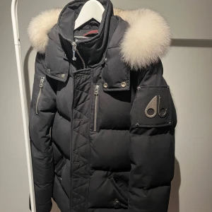 Svart dunjacka med pälsdetaljer - Säljer en snygg svart dunjacka från Moose Knuckles med fluffig vit päls på huvan. Köpt på NK i Göteborg kvitto finns!