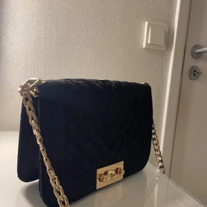 Svart quiltad axelväska med guldkedja - Snygg svart quiltad axelväska med en elegant guldkedja och ett stilrent lås framtill. Perfekt för både vardag och fest. Väskan har en klassisk design som passar till alla outfits.