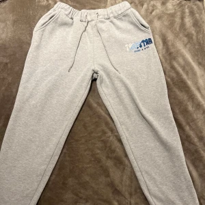 Grå mjukisbyxor med dragkedja - Säljer ett par grå mjukisbyxor i mycket bra skick. De har en elastisk midja med snörning och coola dragkedjor vid bensluten. Perfekta för en chill dag hemma eller en snabb tur till affären. Storleken är inte angiven men de ser ut att ha en normal passform. Märket är inte helt tydligt men det står något i blått och vitt på sidan.