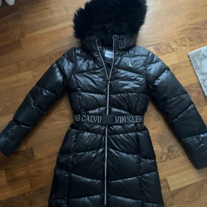 Svart dunjacka från Calvin Klein - Säljer en snygg svart dunjacka från Calvin Klein. Jackan har en fluffig huva och ett bälte med logga i midjan. Den är perfekt för kalla vinterdagar och har en dragkedja framtill. Jackan är i nyskick och håller dig varm och stilren hela säsongen.