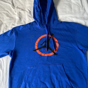 Blå Air Jordan hoodie - Säljer en snygg blå Air Jordan hoodie med det klassiska Jumpman-loggan i svart och orange på bröstet. Hoodien har en stor ficka fram och justerbara snören med orange detaljer. Perfekt för både vardag och träning! Priset går att diskuteras vid snabbt köp