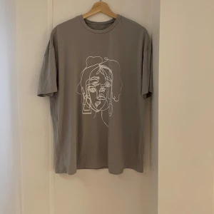 Grå t-shirt med konstnärligt tryck - Säljer en stilren grön t-shirt med ett coolt konstnärligt tryck i vitt på framsidan. T-shirten är oversize och är kortärmad, perfekt för vardagsbruk. Materialet känns mjukt och bekvämt, vilket gör den idealisk för både avslappnade och kreativa tillfällen.