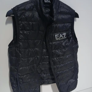 Svart dunväst från Emporio Armani - Säljer en snygg svart dunväst från Emporio Armani EA7. Västen har en quiltad design och en dragkedja framtill. Perfekt för höst och vår när du vill hålla dig varm men ändå se stilren ut. Passar både till vardags och lite finare tillfällen.